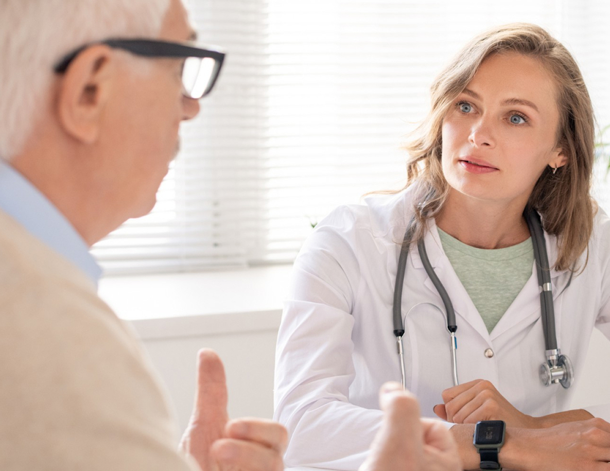 Ärztin im Gespräch mit einem älteren Patienten in einer Praxis; sie hört aufmerksam zu und erklärt etwas.